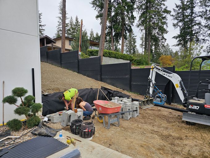 Two people constructing a retaining wall with blocks, hillside backdrop, small excavator.