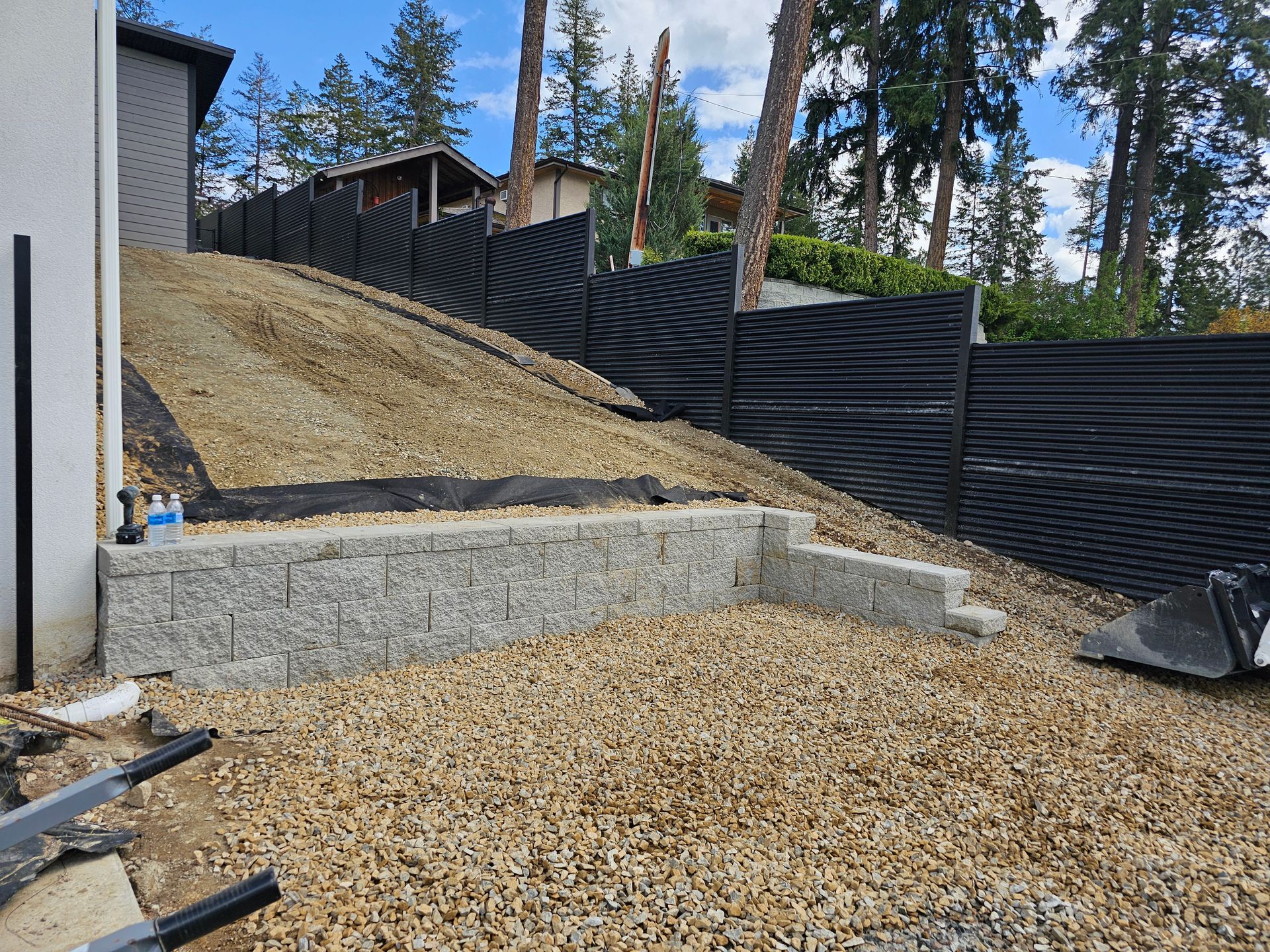 Granite retaining wall and gravel driveway next to a house with a black fence in a hilly setting.