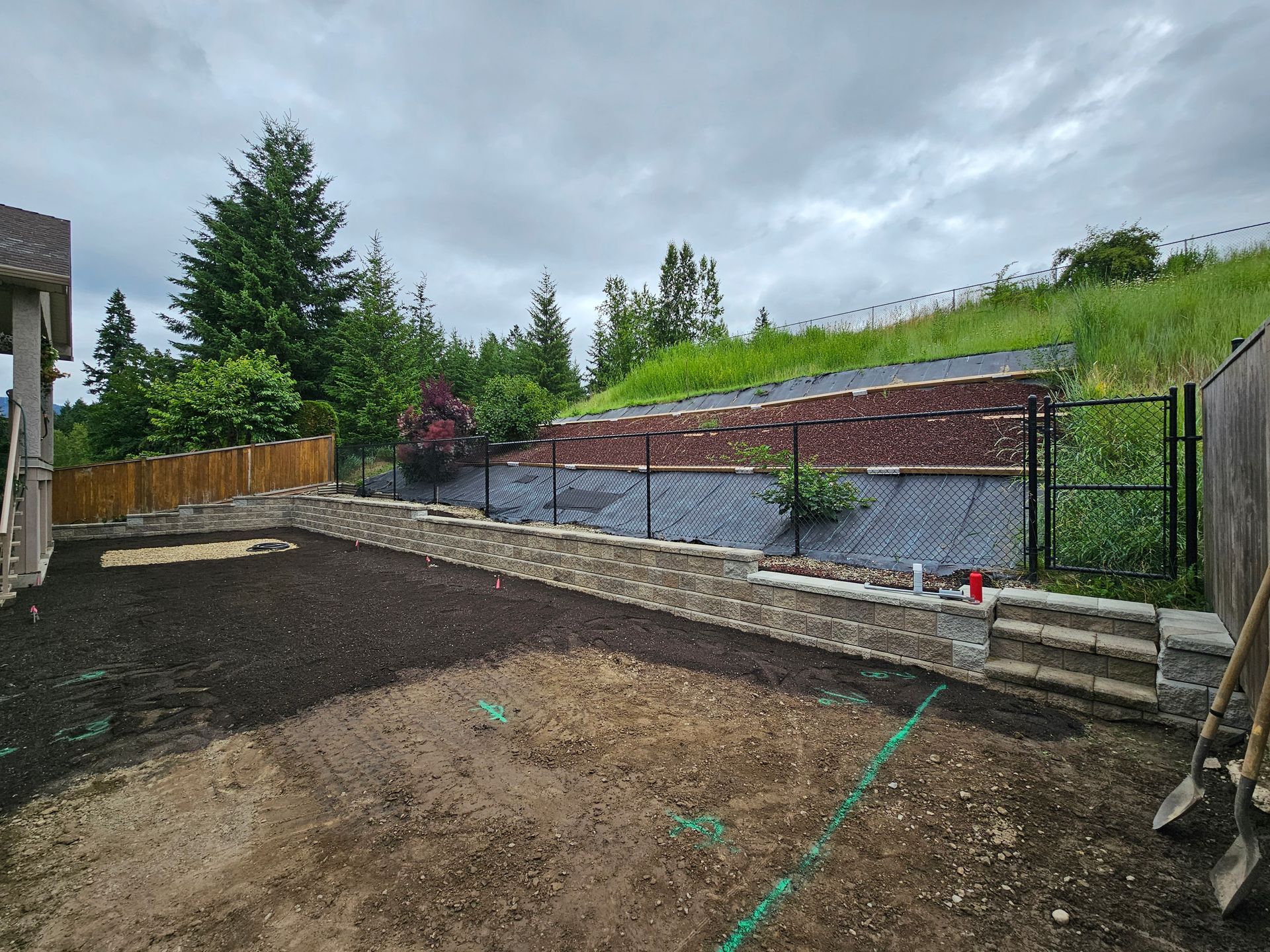 A newly landscaped backyard with a stone retaining wall, fence, and a grassy hill under a cloudy sky.