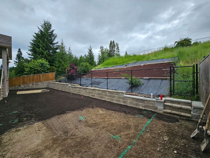 A newly landscaped backyard with a stone retaining wall, fence, and a grassy hill under a cloudy sky.