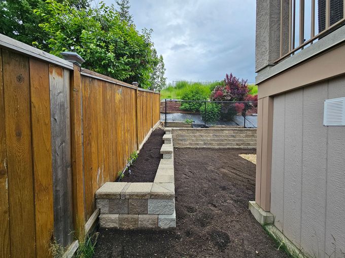 A wooden fence and stone retaining wall border a freshly mulched backyard garden bed next to a beige house.