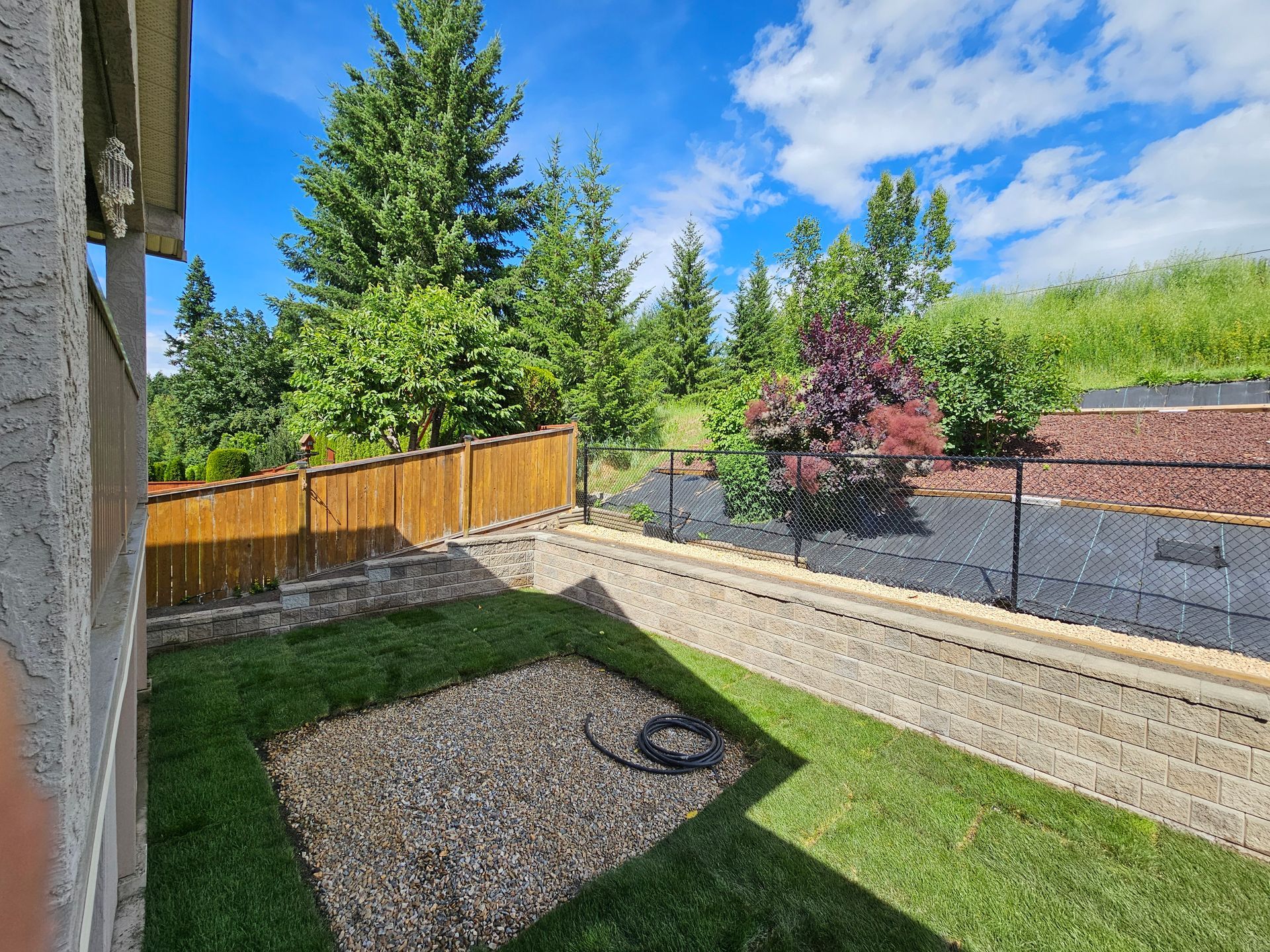 Backyard with retaining walls, grass, rocks, fence, trees, and blue sky.