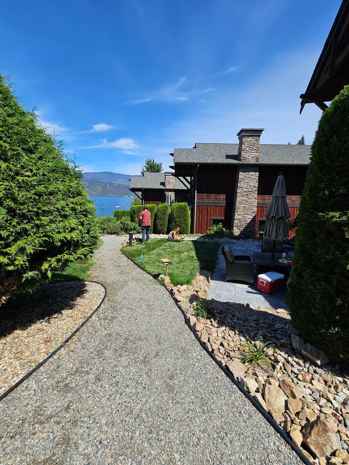 Gravel path leads past a house with dark wood siding and stone chimney to a view of a lake and mountains. Sunny day.