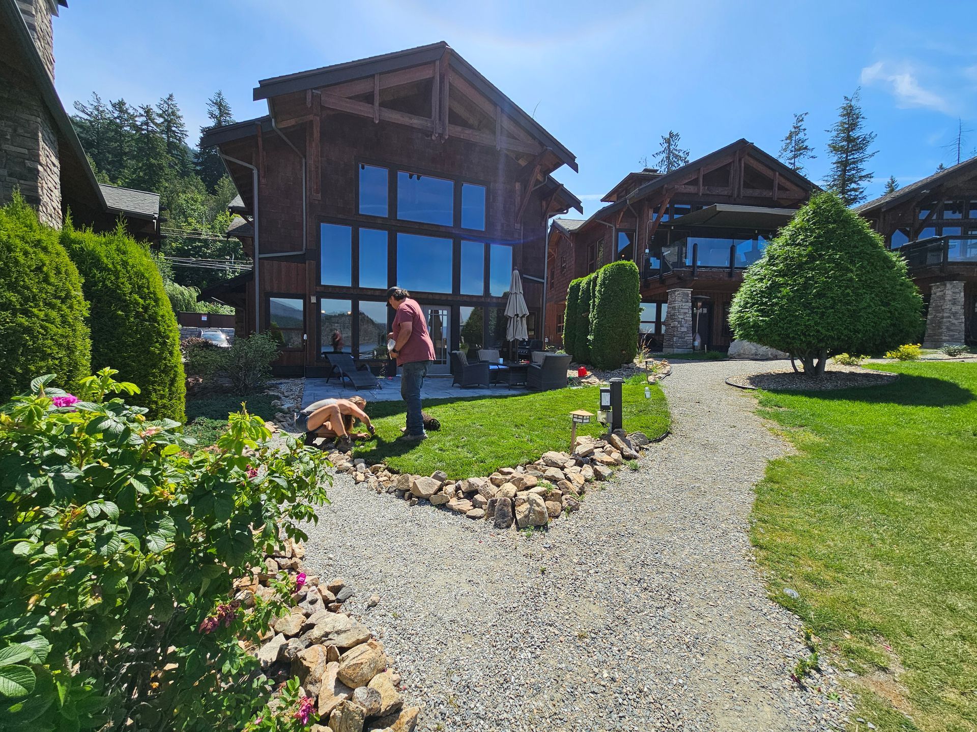 Man tending a garden with a gravel path, surrounded by wooden houses with large windows, bright blue sky.