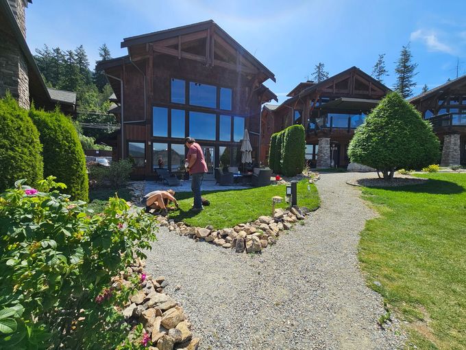 Man tending a garden with a gravel path, surrounded by wooden houses with large windows, bright blue sky.