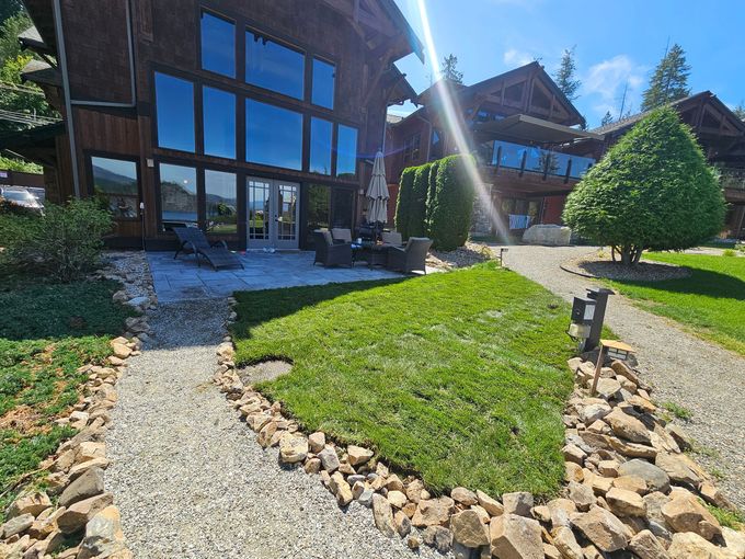Large wooden cabin with big windows, patio, and green lawn. Gravel pathway and a manicured tree are present.