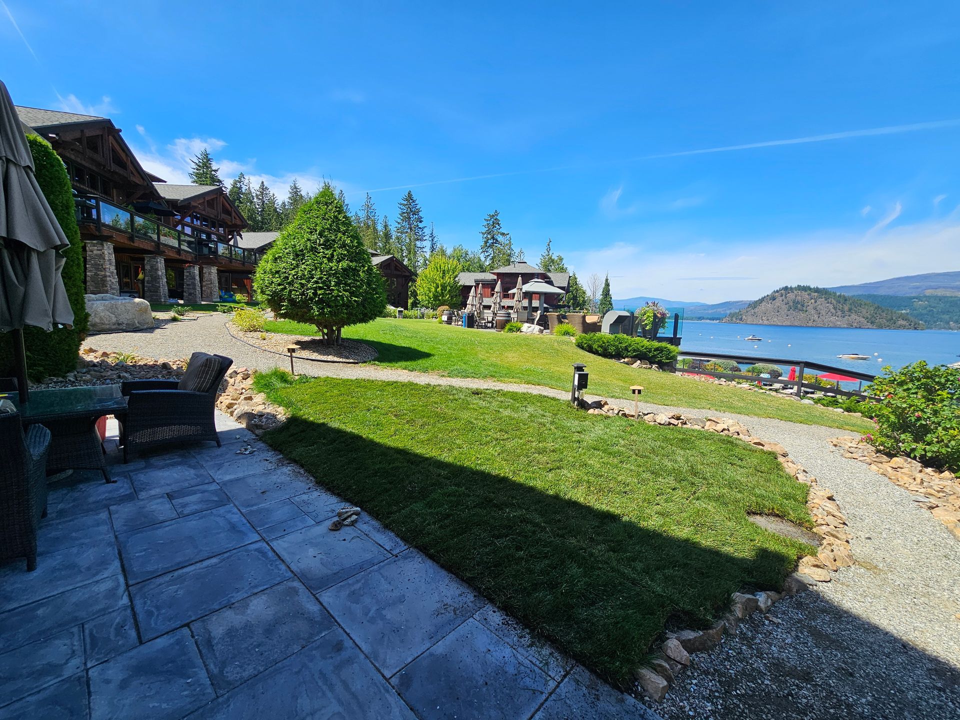 Lakeside resort with green lawn, patio, and buildings. Blue sky, lake, and distant mountains.