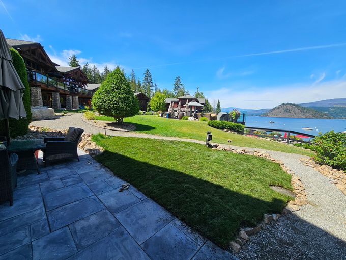 Lakeside resort with green lawn, patio, and buildings. Blue sky, lake, and distant mountains.