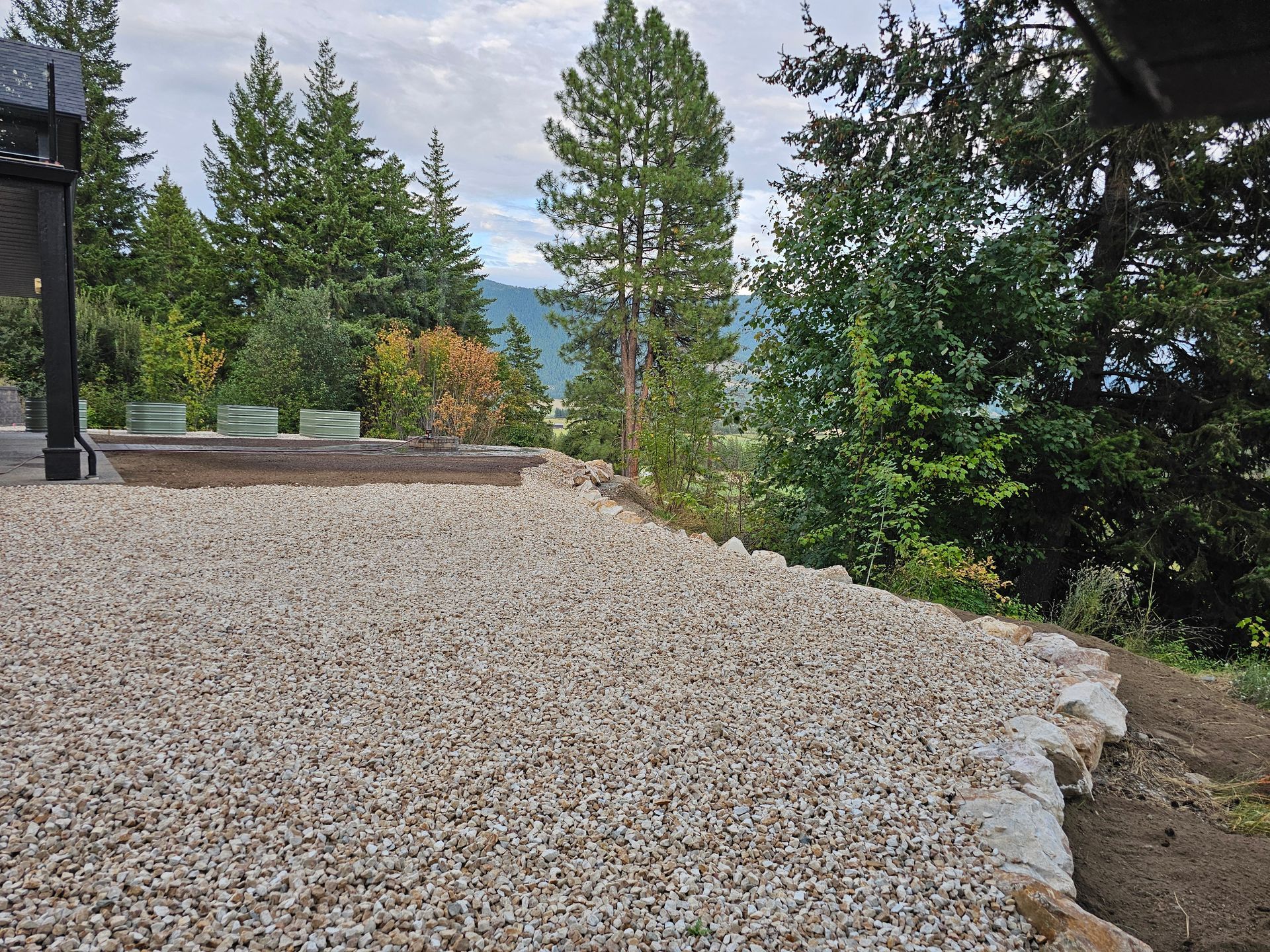 Gravel patio with stone edging overlooks a forest and mountains, cloudy sky.