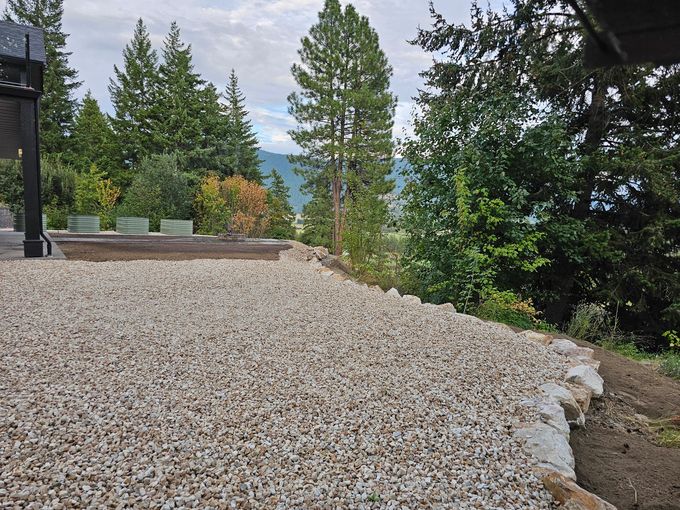 Gravel patio with stone edging overlooks a forest and mountains, cloudy sky.