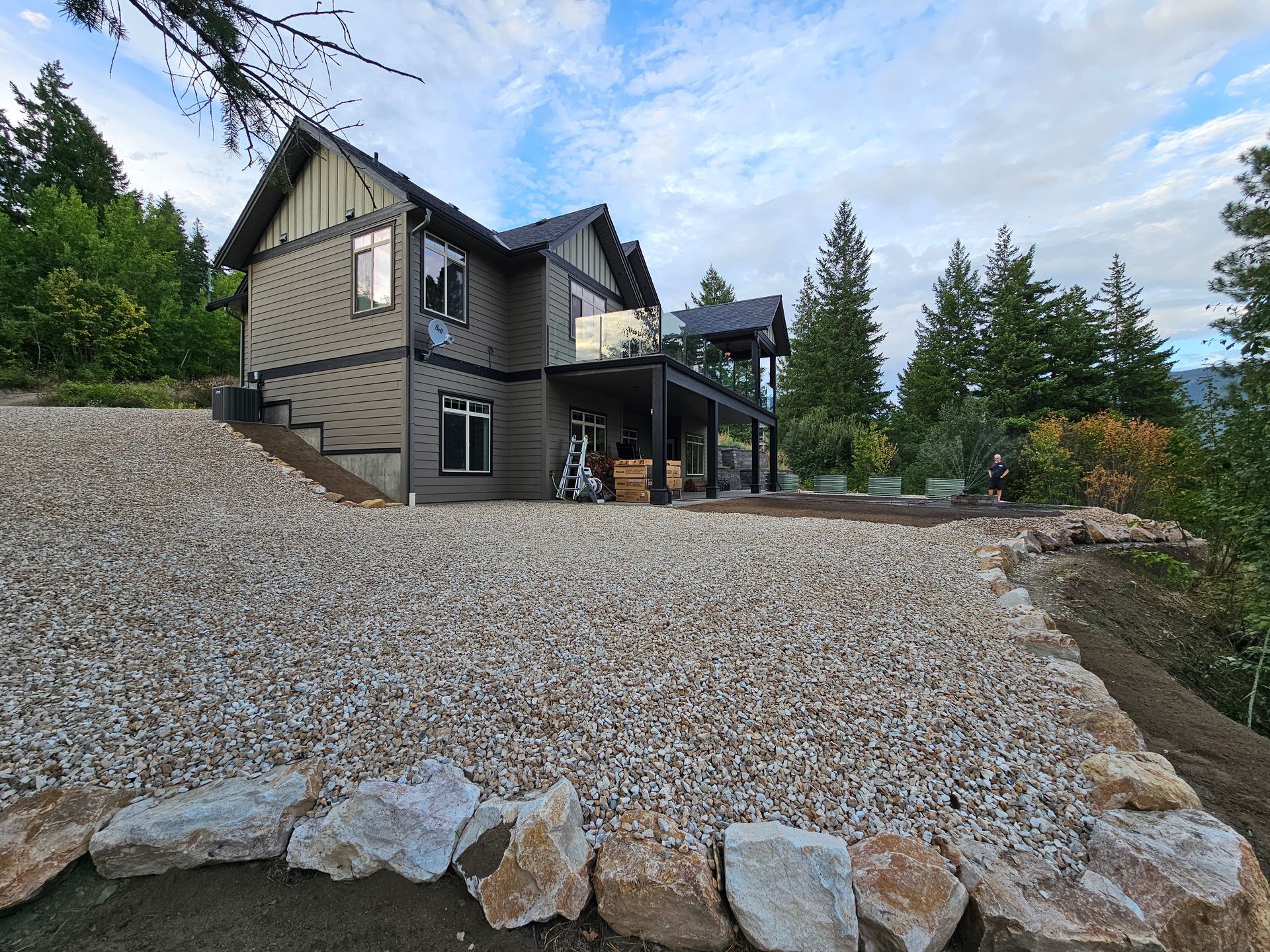 Two-story house with gray siding and gravel landscaping, set against a backdrop of trees and a cloudy sky.