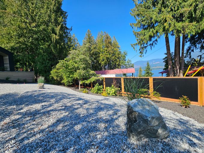 Gravel yard with a black-fenced garden, trees, and a lake view on a sunny day.