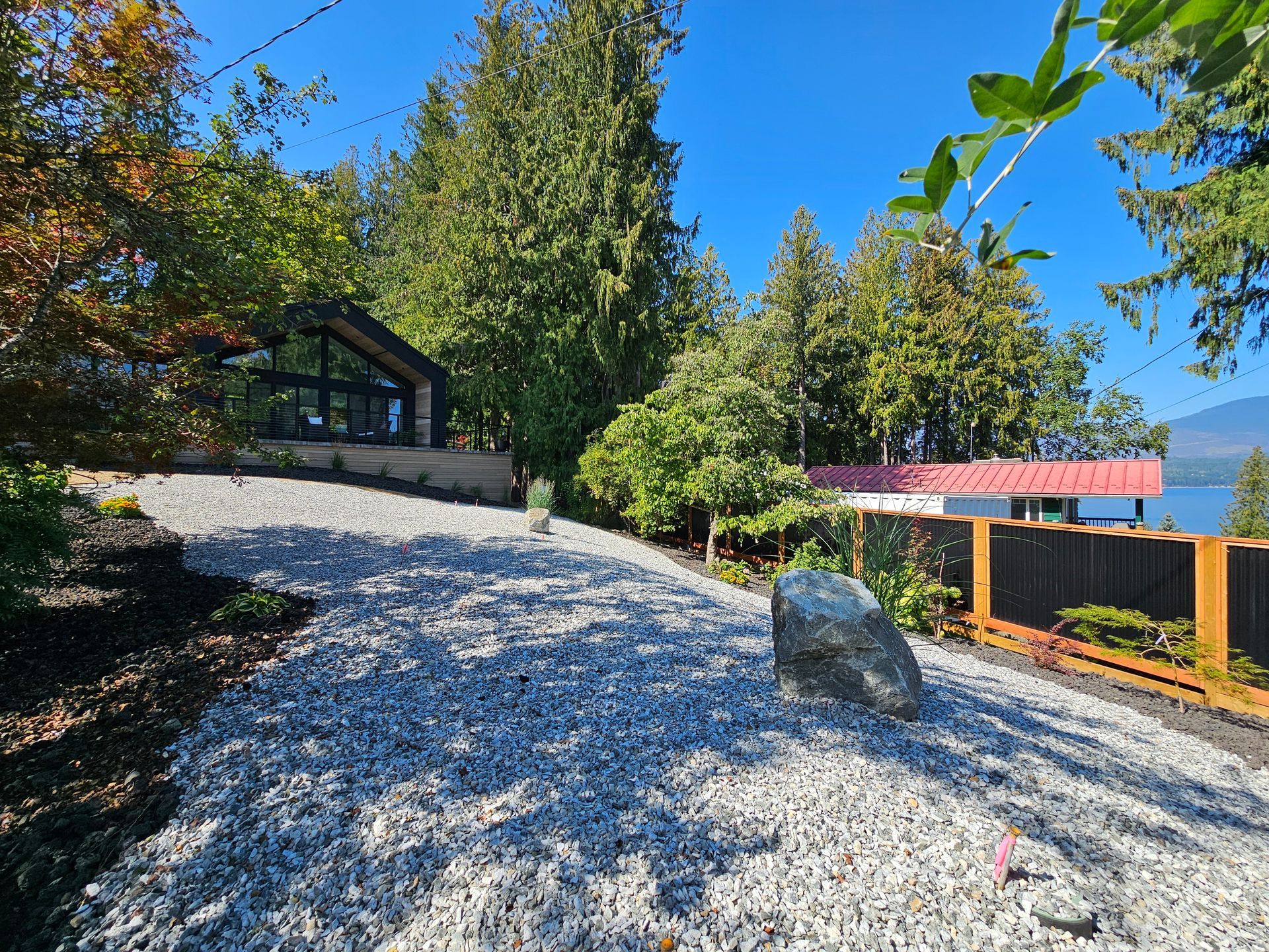 Gravel driveway slopes up towards a modern house with a large window. A large boulder sits on the side.