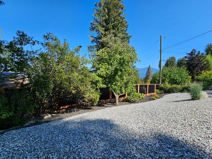 Gravel driveway leads to landscaped yard with trees and a wooden structure; mountains in the background under blue sky.