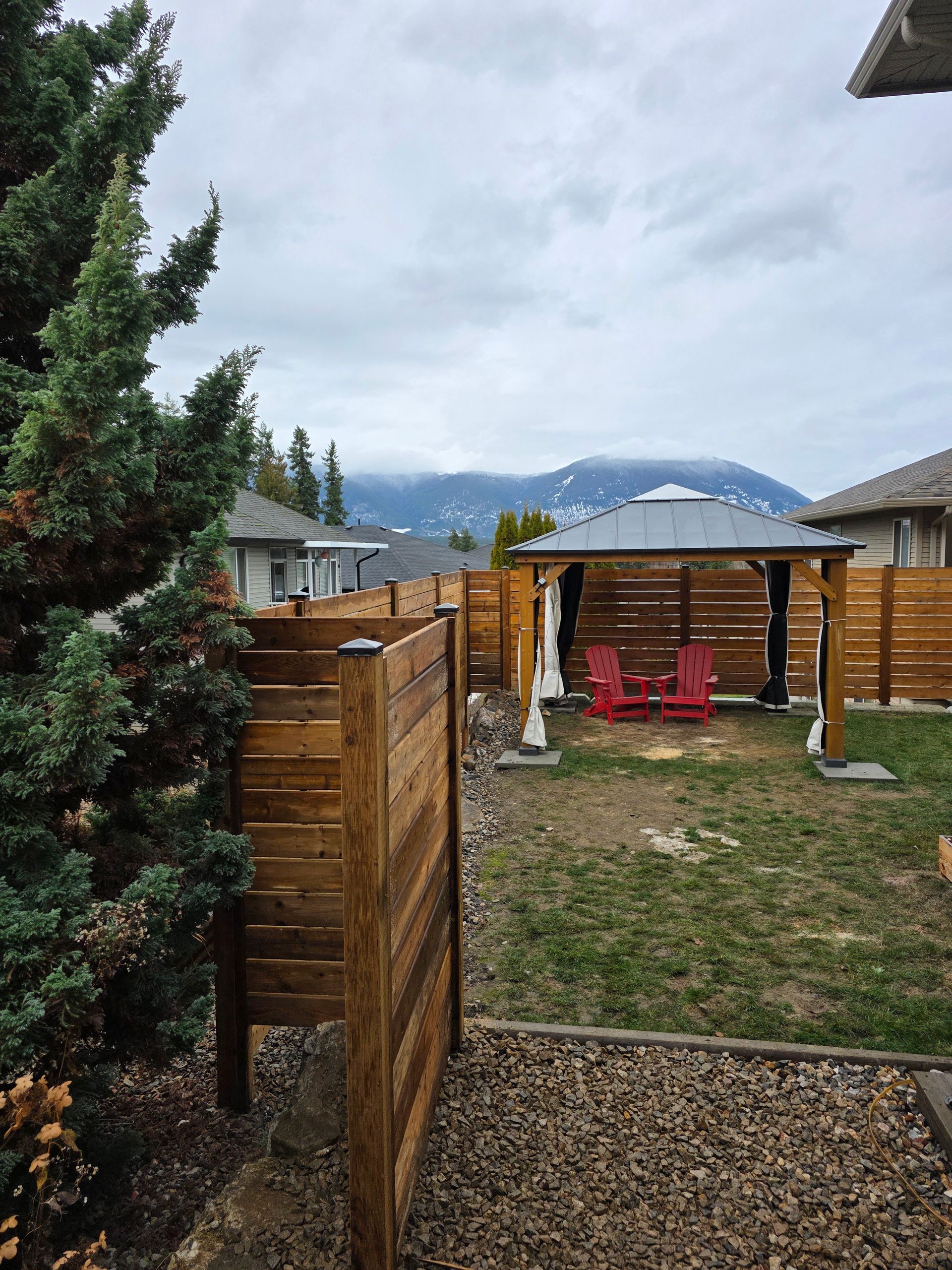 Backyard with a wooden fence, gazebo with red chairs, and a mountain in the background under a cloudy sky.