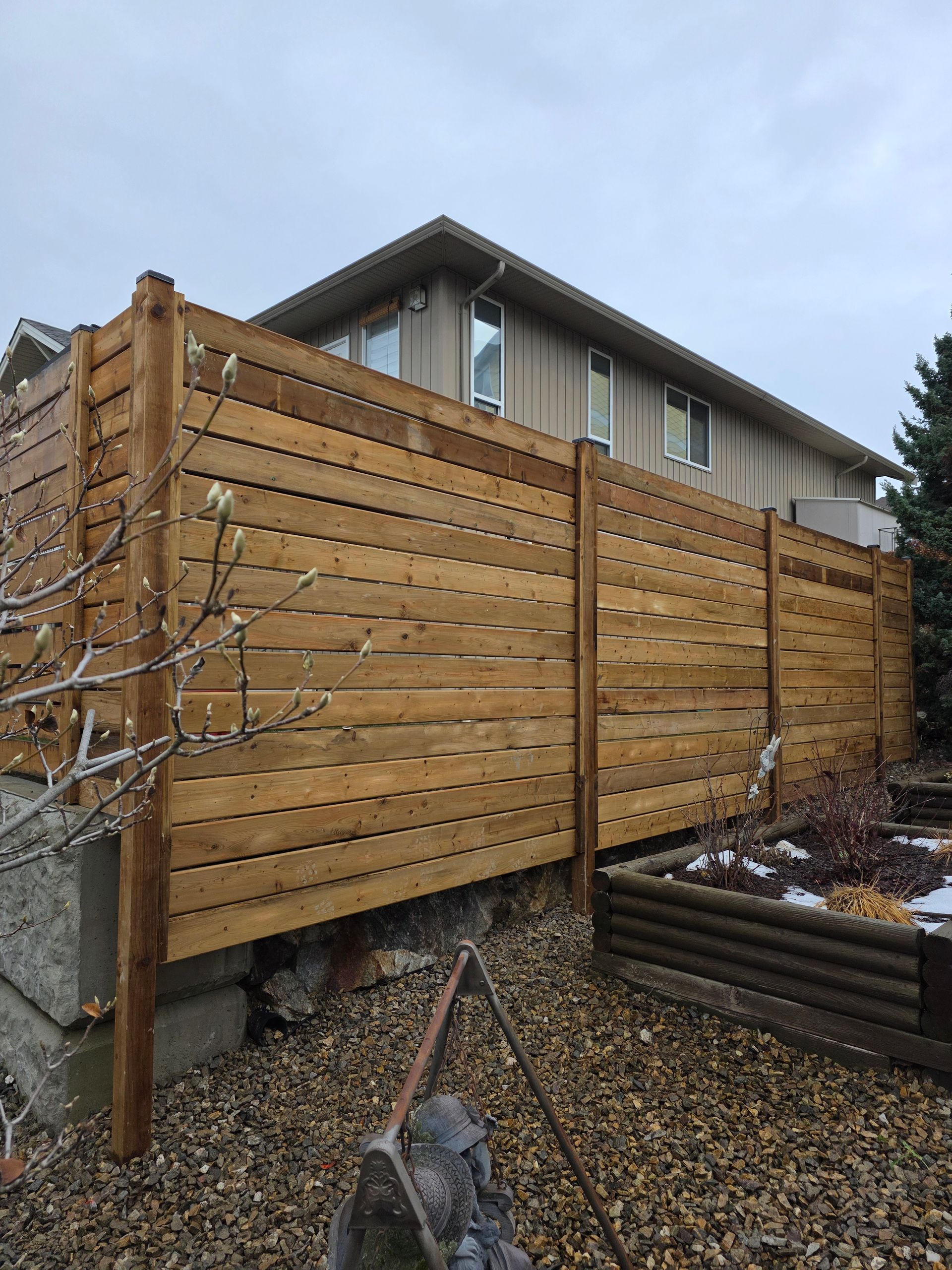 Wooden fence along a house, built on a concrete foundation, with a cloudy sky.