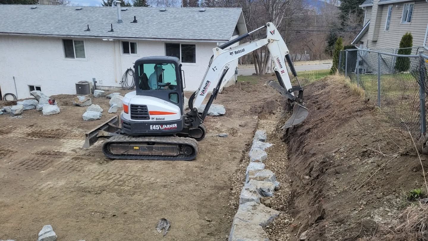A small Bobcat excavator digs in a yard next to a stone retaining wall.