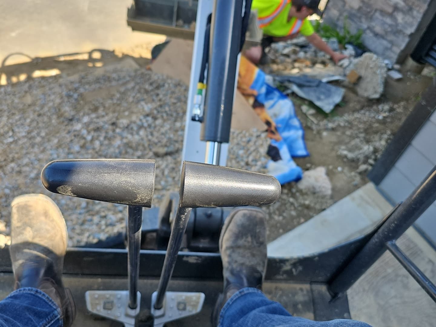 View from inside excavator cab with levers in view, a worker outside.