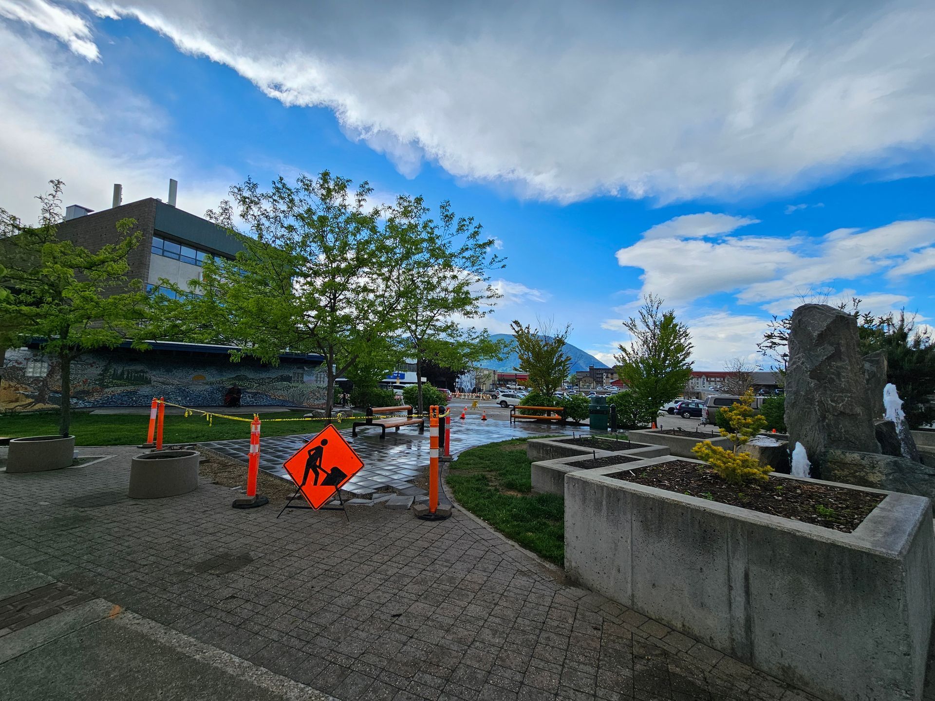 An outdoor construction zone with orange caution signs and barriers in a landscaped park area under a bright, cloudy sky.