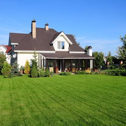 Large two-story house with a brown roof and a porch, set in a well-manicured green lawn under a blue sky.
