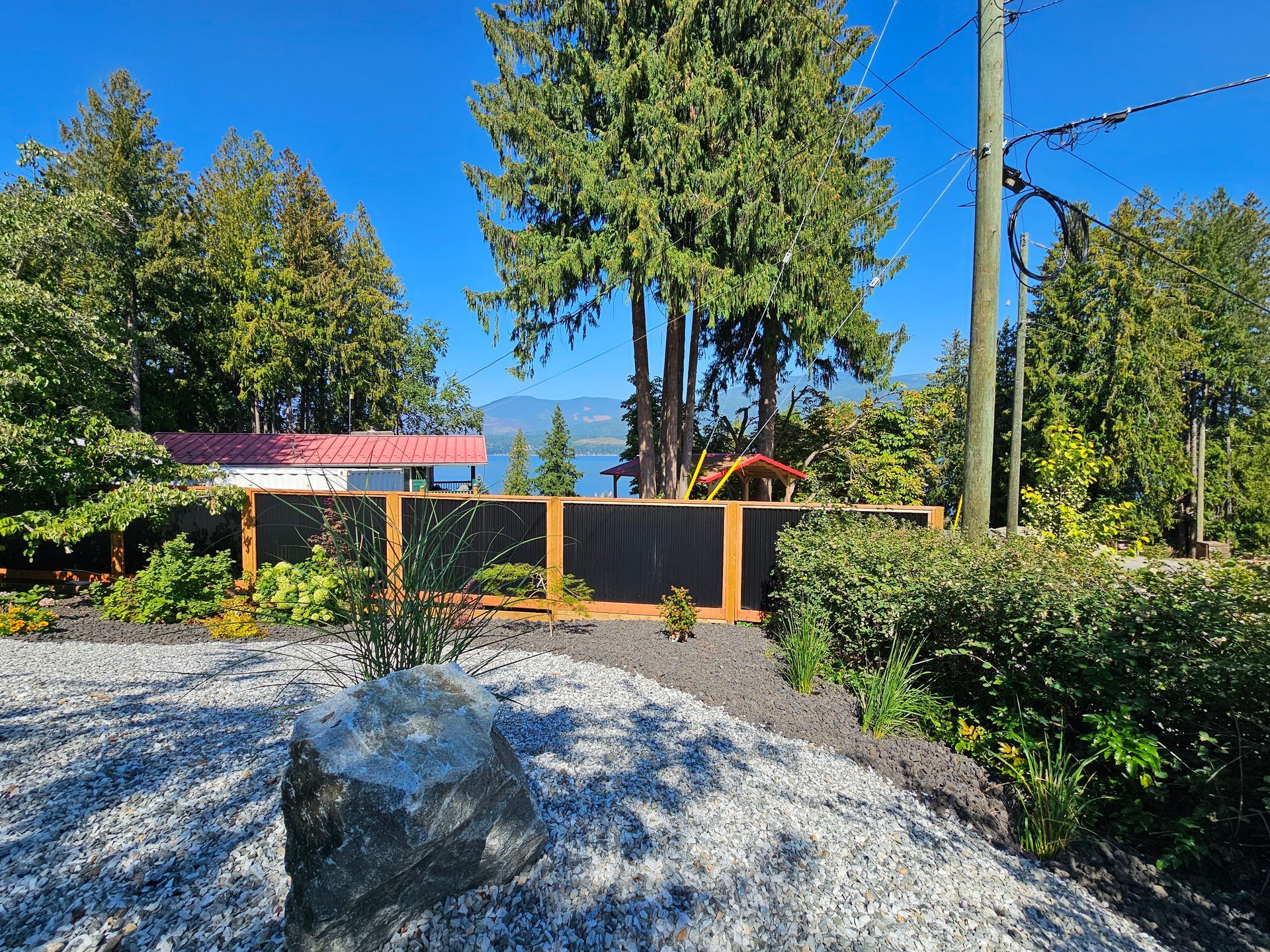 A wooden fence with dark screens sits in a landscaped yard with gray gravel, green bushes, tall trees, and a distant lake.