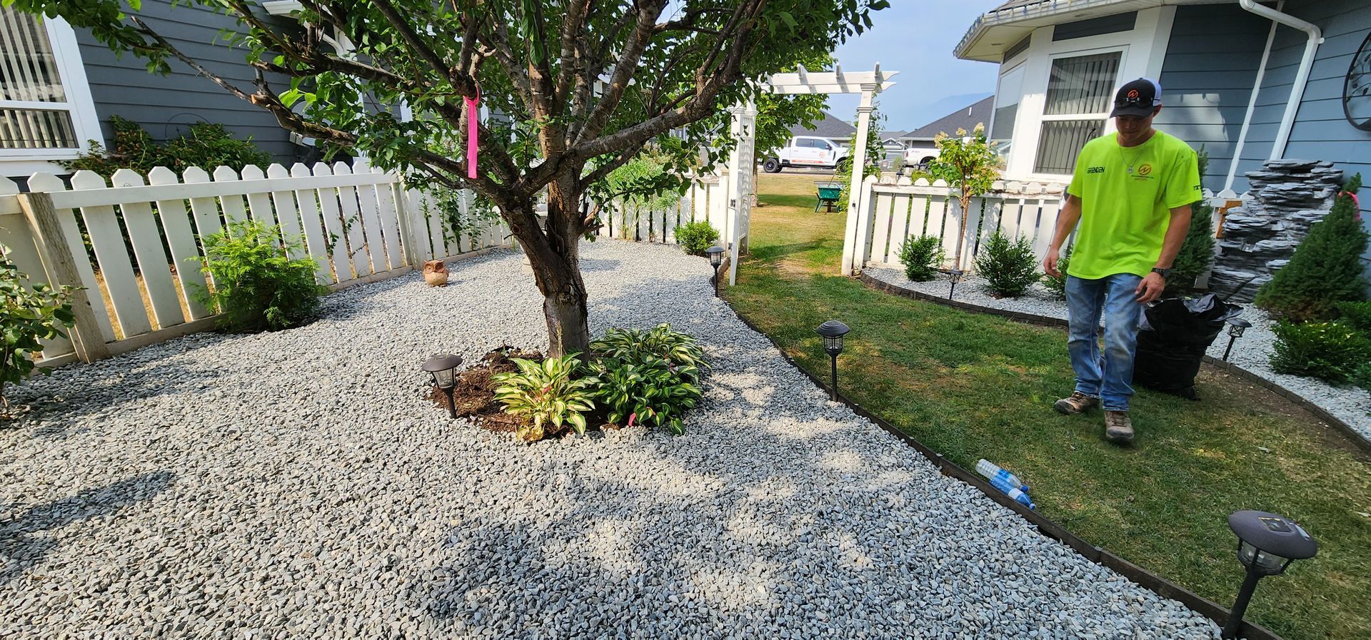 A man in a neon green shirt walks on a grassy path next to a stone-covered yard with a tree.