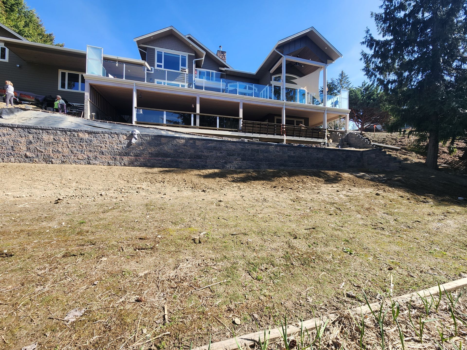 A large, modern house with a wooden deck and glass railing, situated above a stone retaining wall and a grassy yard.