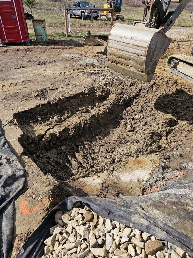 Excavator digging trench in dirt on construction site; rocks in a black liner in foreground.