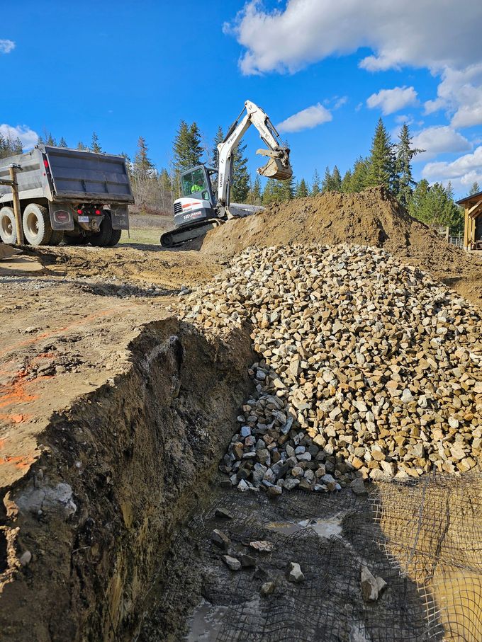Construction site: Excavator, dump truck, dirt pile, and rocks. Blue sky with trees in the background.