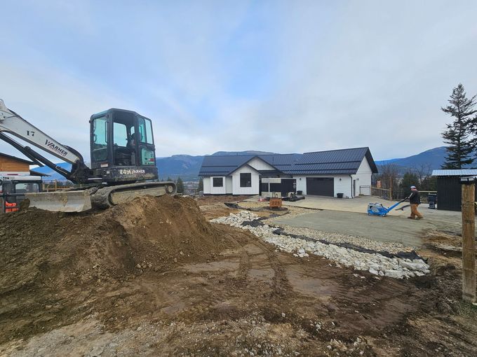Construction site with excavator and house; dirt pile in foreground, gravel base in middle, house in background.