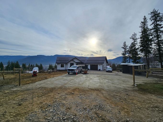 House with vehicles in driveway, mountains and trees in the background under a cloudy sky.