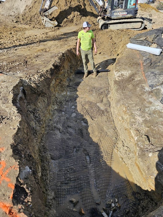 Construction worker standing in a deep trench, with a small excavator in the background.