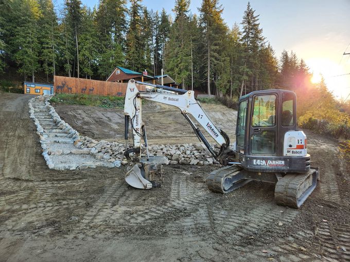 A Bobcat excavator on a hillside, grading soil and moving rocks for construction, with trees in the background.