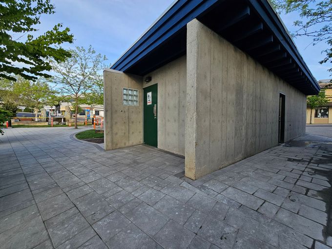 Public restroom building with concrete walls, green door, and blue roof. Paved walkway and trees in background.