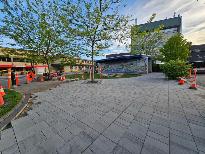 Workers by construction zone with orange cones in a plaza with gray brick pavers and mural, trees, and building.