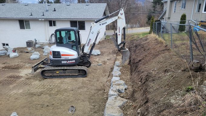A Bobcat excavator digging a trench next to a retaining wall in a backyard.