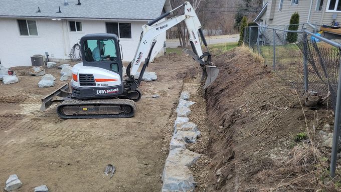 A small Bobcat excavator digging a trench next to a rock wall in a yard.