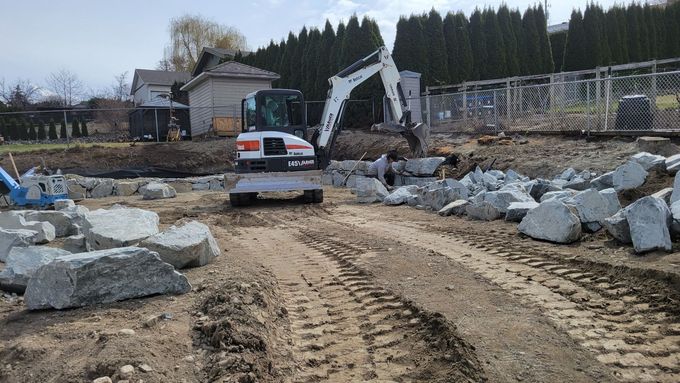 A Bobcat excavator moving large gray rocks in a construction site with a backyard and trees.
