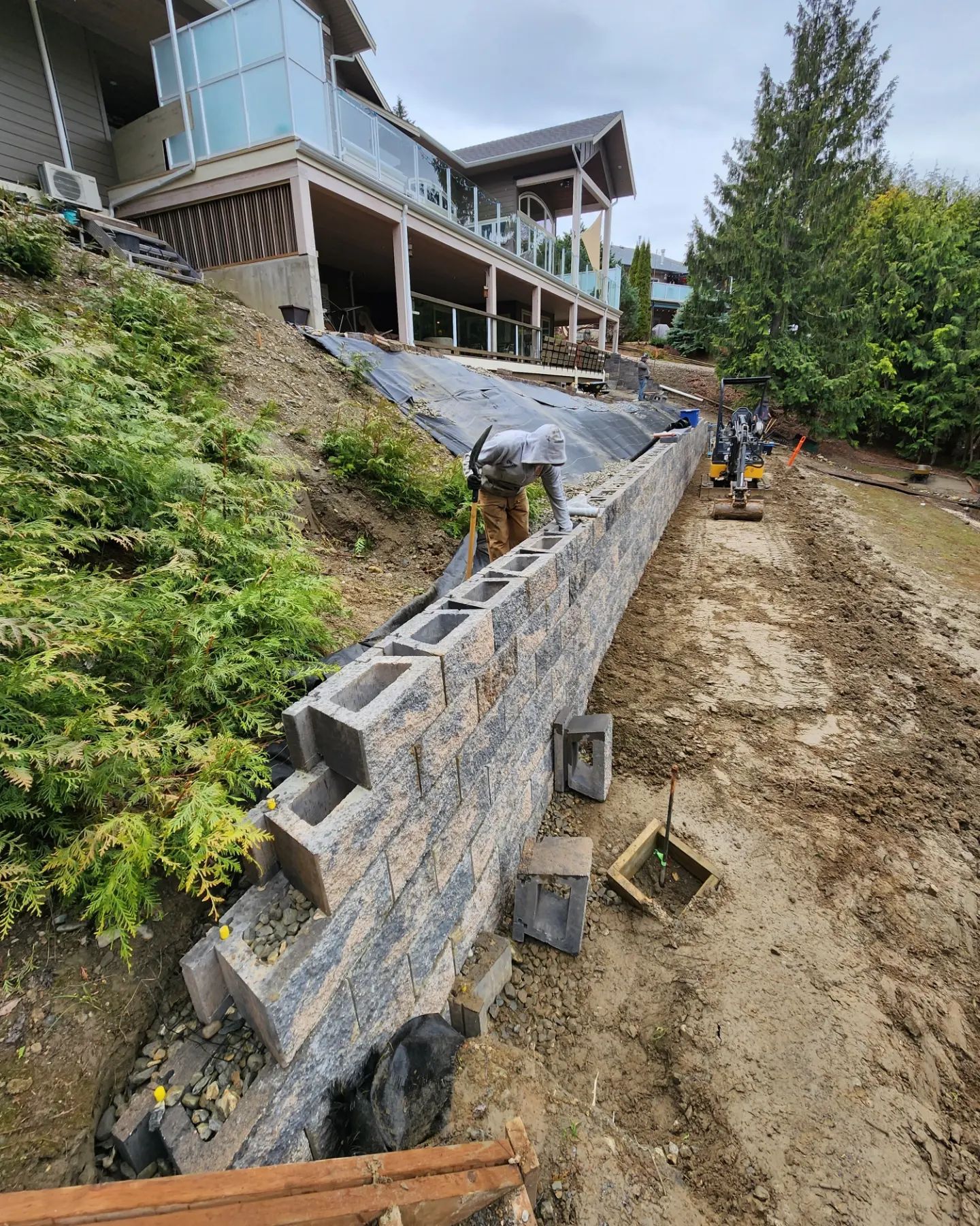 Construction worker building a retaining wall on a hillside near a house. Grey and brown blocks.