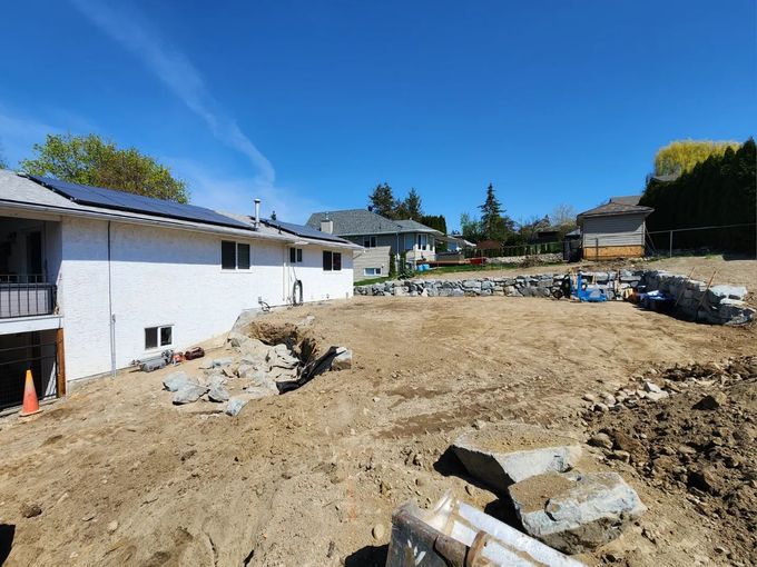 Excavated backyard with dirt, debris, and retaining wall; house with solar panels and blue sky.