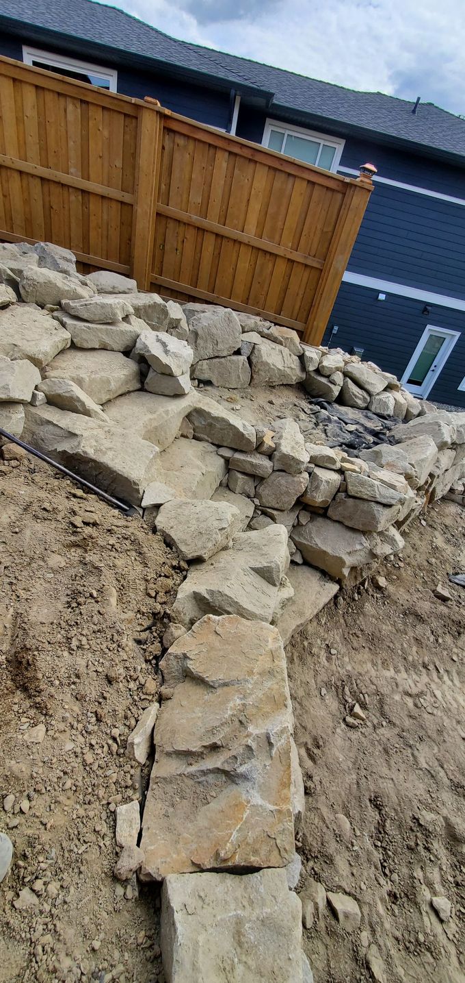 A retaining wall made of stacked stones and soil. A wooden fence and a blue house are visible.