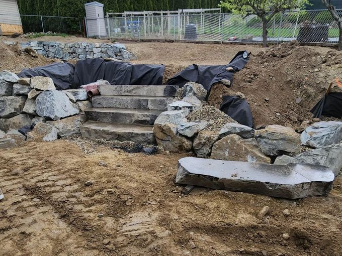 Stone steps under construction, with a stone wall, dirt, and black tarp, outdoors.