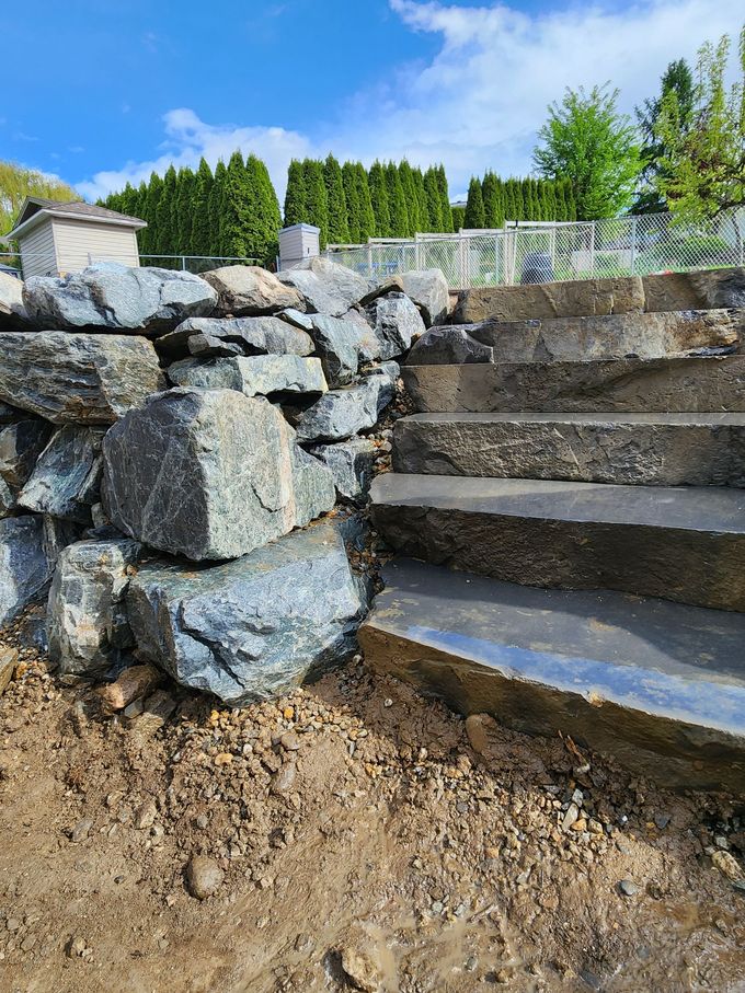 Stone retaining wall beside concrete steps. Sunlight, blue sky, green trees.