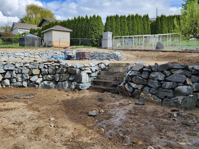 Stone retaining walls with steps, newly cleared dirt, green trees and a blue sky.