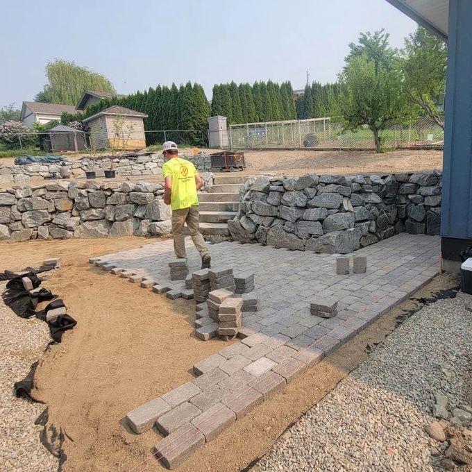 Man laying brick pavers in a backyard patio with stone walls, sandy ground, and a blue house.