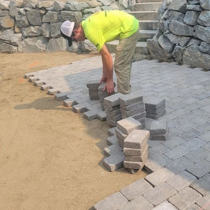 Man laying paving stones on a sandy surface; a stone wall and stairs are in the background.
