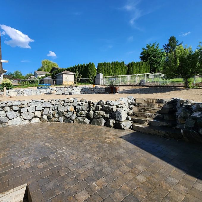 Stone patio with retaining wall, steps, and backyard under a blue sky.