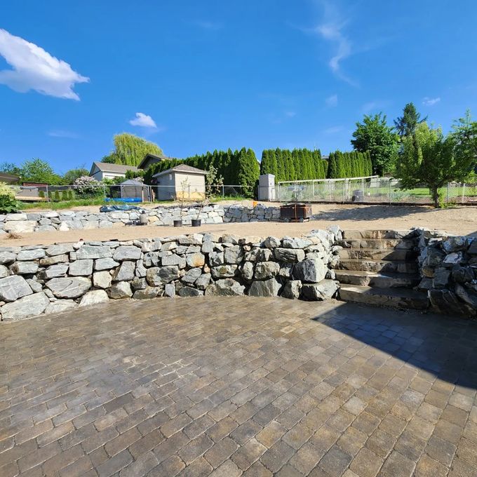 Stone retaining walls and a paved patio in a yard on a sunny day.