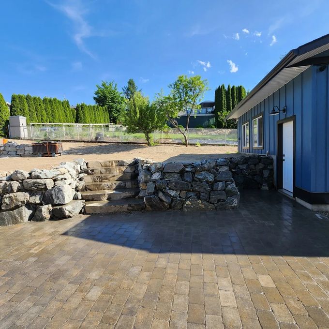 Stone patio with blue building, stone retaining wall, and steps under a sunny sky.