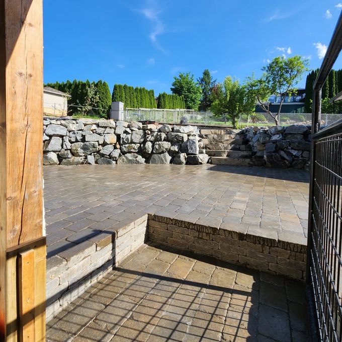 Stone patio with stone wall, steps, and blue sky. A fence on the right, and a wooden post on the left.